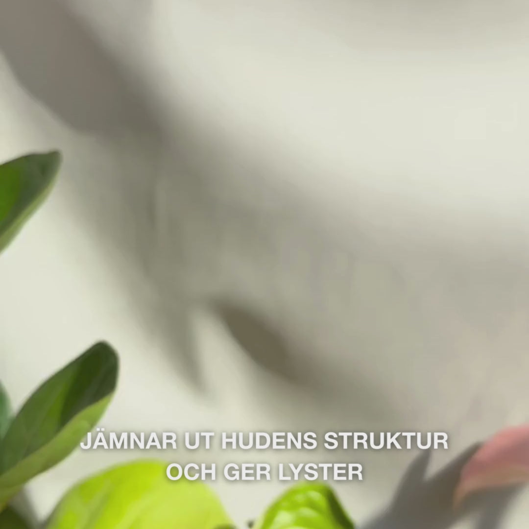 Närbild av en hand som doserar ett par tunna droppar av ett transparent, lätt peeling‑serum på fingertopparna och varsamt masserar in dem i ren ansiktshud — dess silkeslena, flytande textur löser synligt döda hudceller, förfinar hudens struktur och ger lyster med naturliga AHA (Lime Pearl), niacinamid och tea tree‑olja 