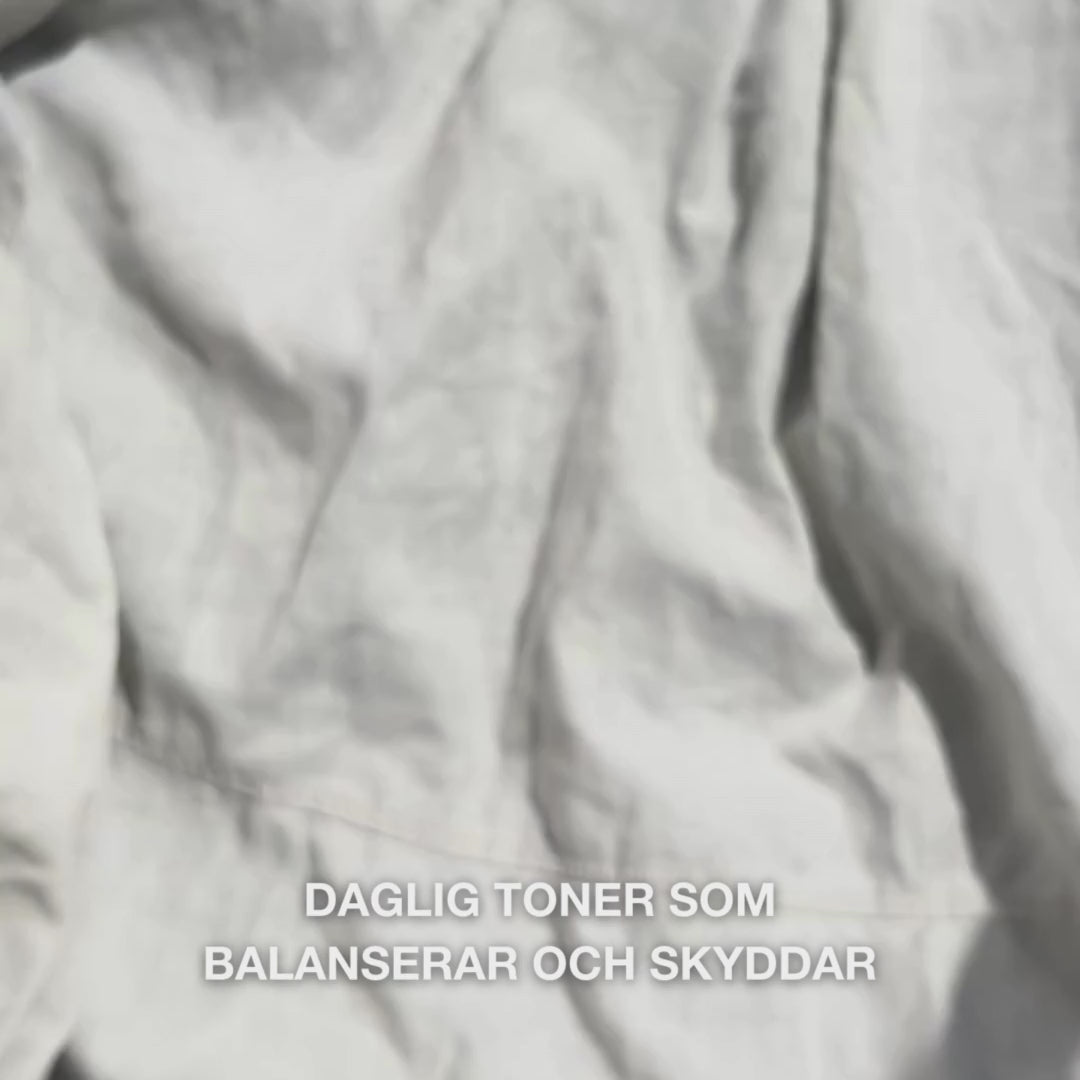 Närbild av en hand som droppar några droppar av ett klart, vattnigt Face Tonic med Tea Tree Oil på en bomullspad och sedan sveper den försiktigt över ren ansiktshud: det lätta, uppfriskande vätskan lugnar, djuprengör, återfuktar och lämnar huden förnyad och redo för ansiktskräm.